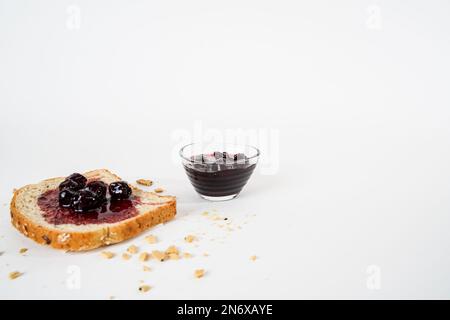 Hausgemachte saure Kirschmarmelade, kleine Schüssel und eine Scheibe Brot mit isolierter Marmelade auf weißem Hintergrund. Stockfoto