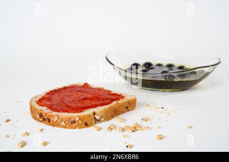 Heiße Tomatensoße auf einer Scheibe Brot und reife schwarze Oliven, mariniert in Olivenöl auf einer Glasplatte, isoliert auf weißem Hintergrund. Stockfoto