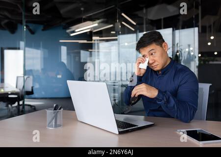 Wässrige Augen eines Mannes im Büro, ein Geschäftsmann übermüdet und überlastet mit Arbeit, verletzt die Augen, ein asiatischer Mann bei der Arbeit mit einem Laptop in der Brille und einem Hemd in der Nähe des Fensters. Stockfoto