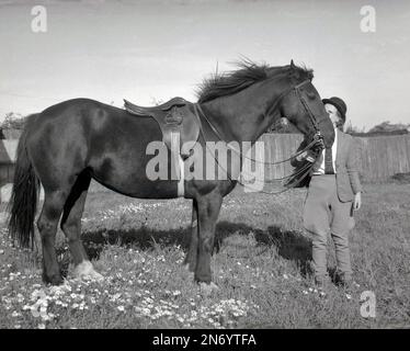 1962, historisches, ländliches Leben....stehend in einer Koppel, eine junge Frau in Cordhose Jodhphurs, Reitjacke & Krawatte und Schutzhelm, die ihrem Pferd einen liebevollen Kuss auf die Stirn gibt, Stetchwork, England, UK. Stockfoto