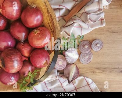 Foto-Komposition mit ganzen Zwiebeln in traditioneller Holzschüssel, Hintergrund mit Holztisch und Zwiebelscheiben, Petersilienzweig, Küchenhandtuch und Stockfoto