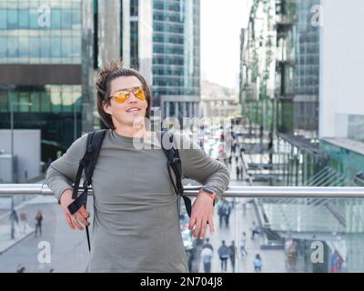 Ein junger Mann mit gelber Brille und langen Haaren steht auf dem Hintergrund einer Großstadt. Moderne Gebäude mit Glaswänden. Millennials Lifestyle. Stockfoto