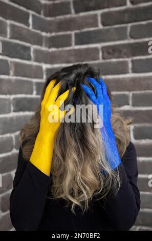 Eine unbekannte Frau, deren Gesicht verzweifelt mit Haaren bedeckt ist, hält den Kopf in den Händen, gemalt in Farben der ukrainischen Flagge. Das Leiden von Zivilisten. Hör Auf Stockfoto