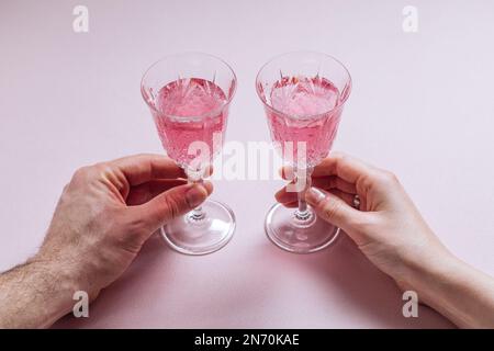 Ein Paar hält zwei rosafarbene Cocktails zum Valentinstag. Liebes- und Jubiläumsatmosphäre mit Herzen. Romantisches Konzept Stockfoto