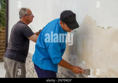 Bild von zwei männlichen Bauarbeitern, die eine Schimmelwand schleifen und reinigen. Problem mit steigender Feuchtigkeit. Stockfoto