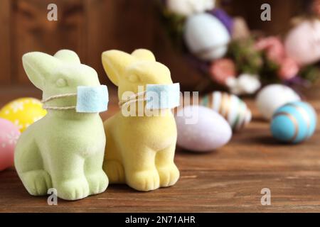 Niedliche Hasenfiguren in Schutzmasken neben gefärbten Eiern auf einem Holztisch, Platz für Text. Osterurlaub während der COVID-19-Quarantäne Stockfoto