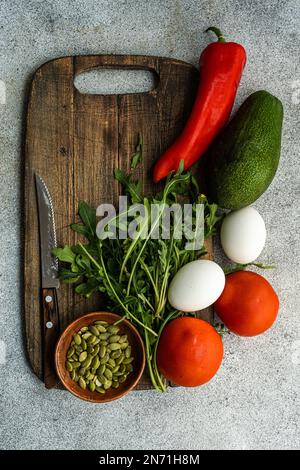 Blick von oben auf hart gekochte Eier, Tomaten, Avocado, Chilipfeffer, Rucola und Kürbiskerne auf einem Holzbrett mit einem Messer Stockfoto