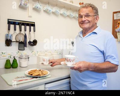 Ruhestand: Tee und Kekse. Ein freundlicher, lächelnder Senior-Mann, der eine englische Tradition aus seiner Küche serviert. Aus einer Reihe von zugehörigen Bildern. Stockfoto