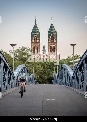 Straßenradfahrer in Freiburg. Auf der "Blauen Brücke", die für den Autoverkehr gesperrt ist. Im Hintergrund befindet sich der Stühlinger Kirchplatz mit den beiden Kirchtürmen der Heiligen Herzkirche. Stockfoto