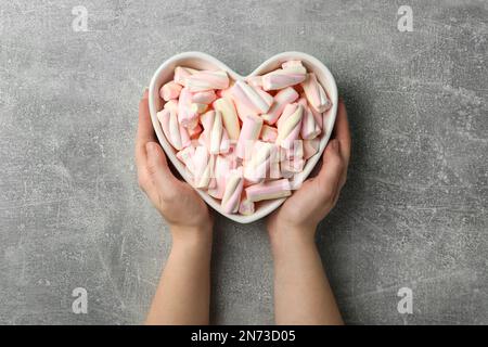 Frau mit herzförmiger Schüssel voller Marshmallows auf grauem Hintergrund, Draufsicht Stockfoto