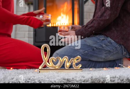 Mann und Frau, die sich zu Hause lieben, sitzen in der Nähe des Kaminherds mit einem brennenden Feuer auf einem gemütlichen Teppich. Valentinstag, glückliches Paar, Liebesgeschichte, Stockfoto