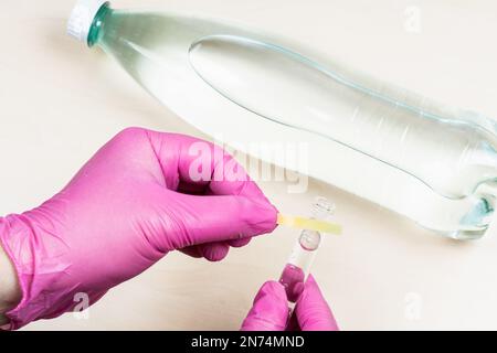 Handschuhe halten feuchtes Lackmuspapier auf dem Reagenzglas, wobei die Probe neutralen Trinkwassers über der Flasche auf dem Lichttisch liegt Stockfoto