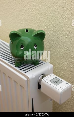 Grünes Sparschwein auf Heizkörper, WLAN-Heizkörperthermostat FRITZ! DECT 302, Display zeigt 1ö °C an, Symbolbild, wird gespeichert, Energie gespart, Energiekosten, steigende Heizkosten, Netzwerke, digitale, Smart-Home-Technologie, Heller Hintergrund, Stockfoto