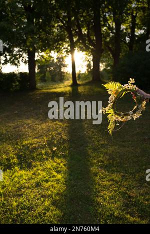 Hand mit einer Blumenkrone während einer Sommerfeier in Lettland. Die Abendsonne scheint durch die Bäume im Hintergrund. Stockfoto