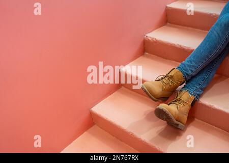 Beine in blauen Jeans braune Stiefel auf rosa Treppen. Werbetexte. Entspanntes Farbkonzept. Stockfoto