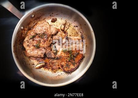 Gebratene Schweinekoteletts mit Kräutern, Gewürzen und Knoblauch in einer Pfanne auf einem schwarzen Kochfeld, Speisen- und Kochkonzept, Kopierbereich, Draufsicht von oben, auswählen Stockfoto