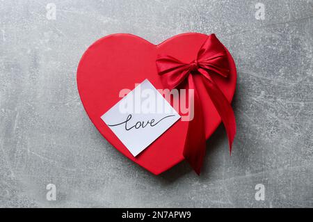 Herzförmige Geschenkbox und Aufkleber mit handgeschriebenem Wort „LIEBE“ auf grauem Hintergrund, Draufsicht Stockfoto