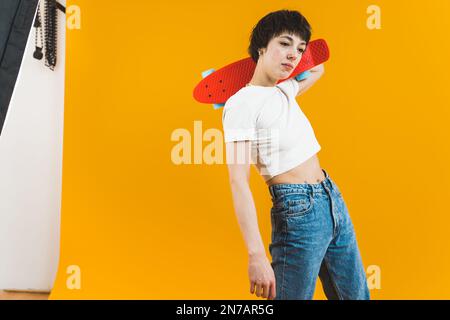 Nicht-binäre Modellpositionierung mit kleinem Skateboard im Studio auf gelbem Hintergrund. Förderung von Sport und Aktivitäten im Freien. Hochwertiges Foto Stockfoto