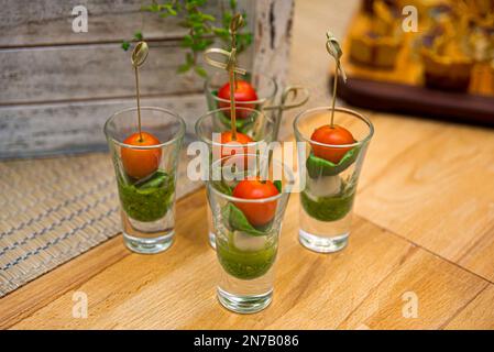 Caprese Canapes in Glasstapeln. Kirschtomaten, Mini-Mozzarella-Bälle, Pesto-Sauce und Basilikumblätter in einem Glas auf einem Holztisch. Stockfoto