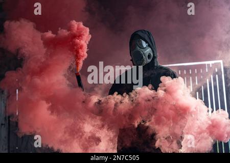 Ein Mann mit Gasmaske, der eine Rauchbombe in der Hand hat. Leuchtend roter, farbenfroher Rauch. Alte tschechoslowakische Gasmaske M-10(M). Stockfoto