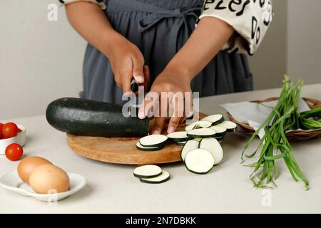 Frau schneidet grünen Zucchini-Kürbis auf einen hellen Holztisch, Kochen Stockfoto