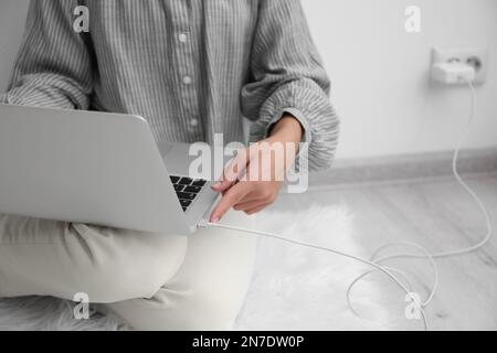 Frau, die das Ladekabel mit dem Laptop verbindet, auf dem Boden in der Nähe der weißen Wand, Nahaufnahme Stockfoto