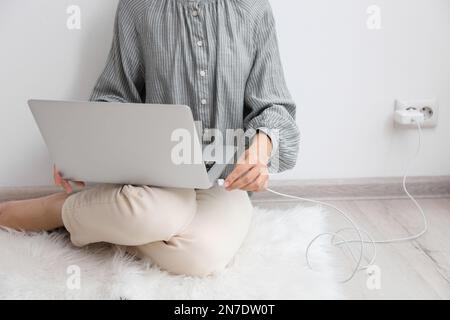 Frau, die das Ladekabel mit dem Laptop verbindet, auf dem Boden in der Nähe der weißen Wand, Nahaufnahme Stockfoto