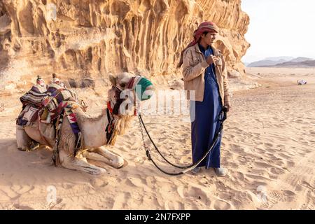 Ein Beduiner gibt an einem sonnigen Tag in der malerischen Landschaft des UNESCO-Weltkulturerbes Wadi Rum vor, mit einem Kamel fotografiert zu werden. Wadi Rum ist bekannt als eine der landschaftlich schönsten Regionen der Welt. Stockfoto