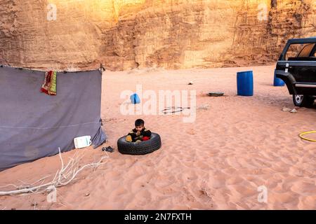 Der Beduinenjunge spielt an einem sonnigen Tag in einem Reifen auf dem Sand in der malerischen Landschaft des UNESCO-Weltkulturerbes Wadi Rum Wüste. Wadi Rum ist bekannt als eine der landschaftlich schönsten Regionen der Welt. Stockfoto