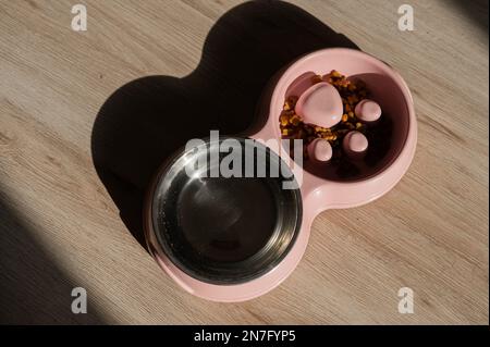 Eine Doppelschüssel für langsames Füttern und eine Schüssel Wasser für den Hund. Draufsicht auf einen rosafarbenen Teller mit trockenen Speisen auf einem Holzboden. Stockfoto