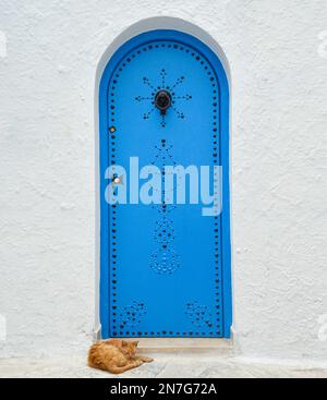 Eine traditionelle blaue Tür mit tunesischen Ornamenten und eine schlafende Katze vor der weißen Wand Stockfoto