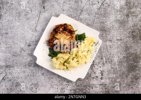 Gebratene Schweinekoteletts mit geschmolzenem Käse und Kartoffelpüree mit Kräutern auf einem weißen quadratischen Teller auf dunklem Hintergrund. Draufsicht, flach liegend Stockfoto