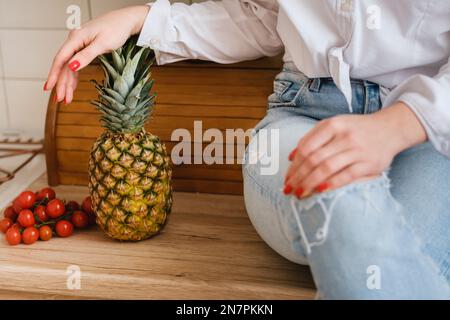 Die Nahaufnahme einer Hausfrau in einem weißen Hemd und Jeans sitzt auf einem Tisch und Obst und Gemüse liegen in der Nähe. Das Konzept der gesunden Ernährung. Stockfoto