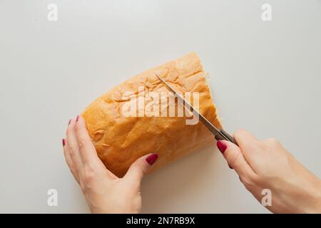 Weibliche Hand schneidet ein Stück Brot mit einem Messer auf einem weißen Tisch in der Küche zu Hause Stockfoto