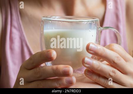 Ein junges ukrainisches Mädchen hält morgens einen durchsichtigen Glasbecher mit Milch Stockfoto