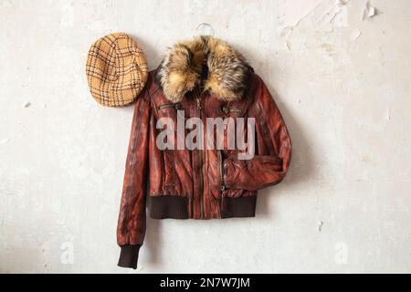 Rote Lederjacke und karierte Mütze, die an der weißen alten Wand hängt, Mode Stockfoto