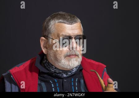 Schönes Porträt von ukrainischen bärtigen älteren schwarzen Brillen, die Tabakpfeifenmann halten, während sie über Lebensprobleme in der Dunkelheit nachdenken Stockfoto