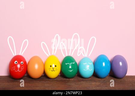 Mehrere Eier mit gezogenen Gesichtern und Ohren, darunter Osterhasen, auf einem Holztisch vor pinkfarbenem Hintergrund Stockfoto