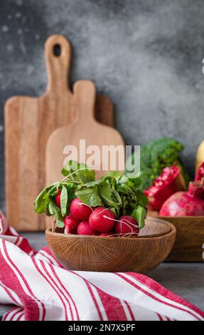 Ein Haufen frischer roher Rettich in einer Holzschüssel auf dem Küchentisch mit frischem Obst, Grün, Gemüse in einem Holztablett auf einem Tisch mit grauem Stein im Hintergrund, kochen Stockfoto