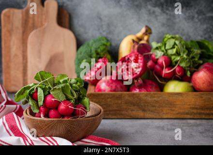 Ein Haufen frischer roher Rettich in einer Holzschüssel auf dem Küchentisch mit frischem Obst, Grün, Gemüse in einem Holztablett auf einem Tisch mit grauem Stein im Hintergrund, kochen Stockfoto