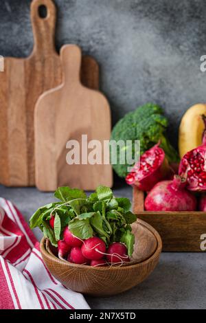 Ein Haufen frischer roher Rettich in einer Holzschüssel auf dem Küchentisch mit frischem Obst, Grün, Gemüse in einem Holztablett auf einem Tisch mit grauem Stein im Hintergrund, kochen Stockfoto