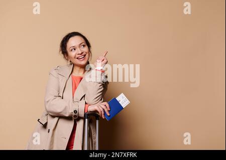 Glückliche, ethnische Passagierin mit Bordkarte in den Händen, lächelt, zeigt mit dem Zeigefinger auf die Textstelle auf beigefarbenem Hintergrund Stockfoto