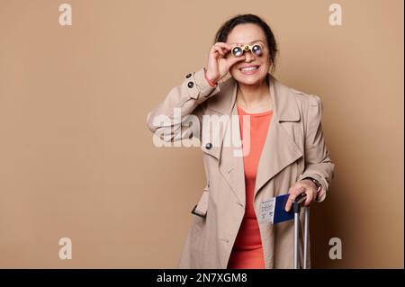 Lächelnde Frau, die durch klassische Operngläser in die Kamera schaut, mit Bordkarte und Koffer auf beigefarbenem Hintergrund Stockfoto