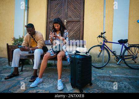 Wunderschöne multiethnische Freunde unterhalten sich auf ihren Telefonen, während sie eine Pause machen und auf einer Bank sitzen Stockfoto