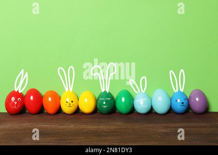 Mehrere Eier mit gezogenen Gesichtern und Ohren wie Osterhasen unter anderem auf einem Holztisch vor grünem Hintergrund Stockfoto