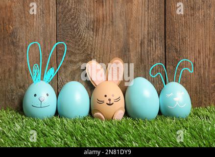 Mehrere Eier mit Ohren als Osterhasen, unter anderem auf grünem Gras vor Holzhintergrund Stockfoto