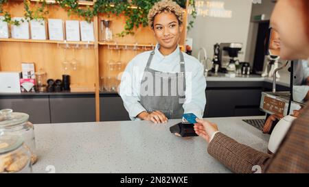 Barkeeper erhält Zahlung von einem Gast. Barista in der Schürze hält POS-Terminal, während der Gast die Purchase Card bezahlt. Stockfoto