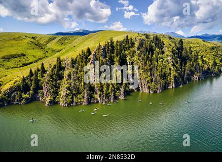 Top-down-Drohnenaufnahme von einer Gruppe von Menschen auf Stand-Up-Paddle-Boards SUP im Bergsee in der Nähe von Felsen mit Fichtenwald in Kasachstan Stockfoto