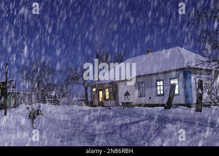Wunderschöner Blick auf das schneebedeckte Haus bei Nacht bei starkem Schneefall. Alles ist mit Schnee bedeckt. Rauch kommt aus dem Kamin. Stockfoto
