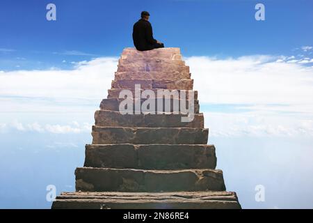 Ein Mann sitzt auf der obersten Stufe einer Steintreppe, die zum Himmel führt. Der Mann blickt auf die Wolken. Stockfoto